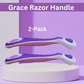 Two Grace Razor Handles in a 2-pack, displayed horizontally on a white cylindrical pedestal against a purple and lavender background. The handles are white with purple textured grips and pink circular accents.